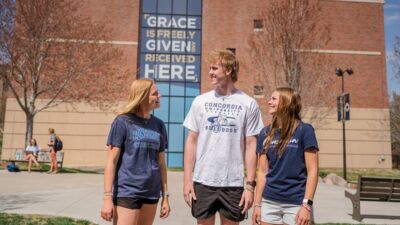 Three students standing outside Thom with the words "Grace is Freely Given and Received Here" in the background.