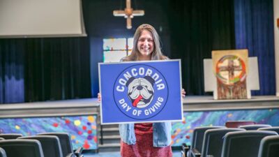 A smiling student in chapel holding up a sign with the Concordia Day of Giving logo on it