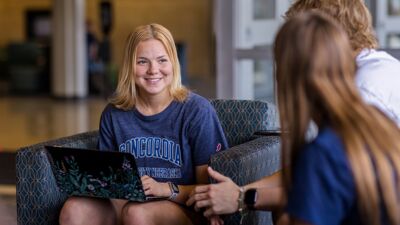 A smiling student sitting with a laptop on her lap, chatting with her two friends