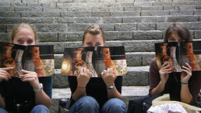 Three students holding magazines in front of their faces, sitting on some steps in Turkey