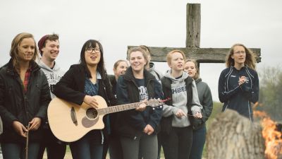 A group of students, on of them has a guitar, standing in front of a cross singing